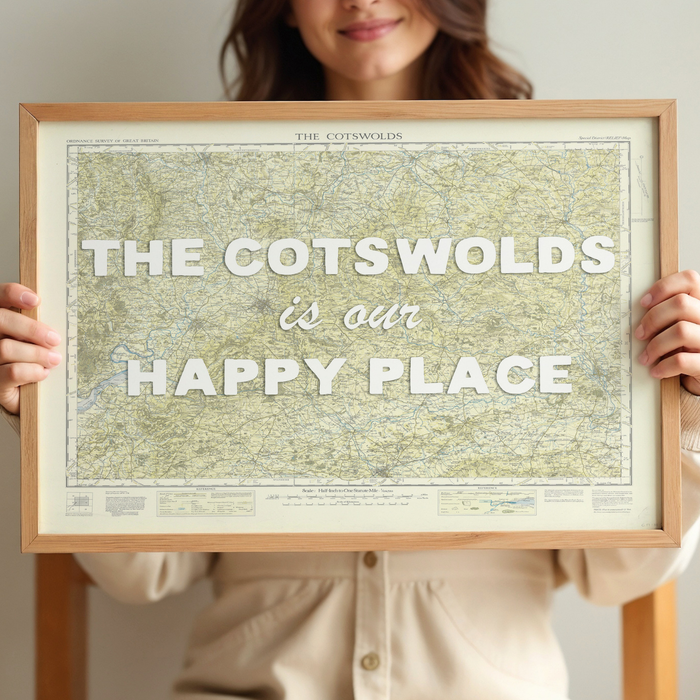 Person holding a framed Cotswolds map with 'The Cotswolds is our happy place' text.