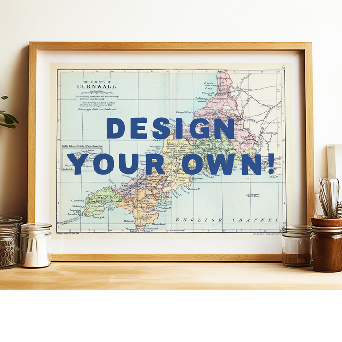 A framed custom map print with the text 'DESIGN YOUR OWN!' displayed prominently over a map of Cornwall, England. The frame is placed on a shelf with various items around it.