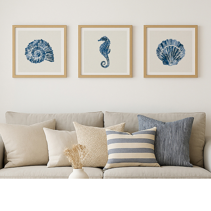 A set of three framed shell and seahorse art prints featuring indigo shell, scallop shell and seahorse artworks, displayed above a beige sofa with various pillows.