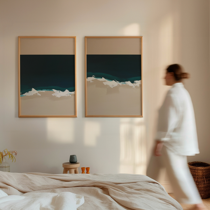 Two framed minimalist coastal artworks on a wall with a person in a white outfit walking by.